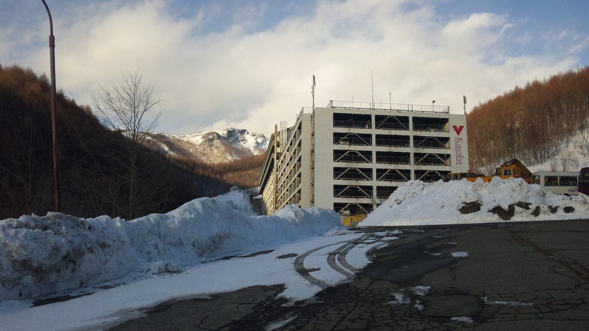 川場滑雪場的立體停車場：多層的白色停車樓，外牆有紅色 V 字標誌和直寫的 Kawaba 字樣，前方道路兩側堆著剷起來的雪，右側停著一輛小巴與黃色工程車，背後是落葉松山坡與覆雪的岩峰