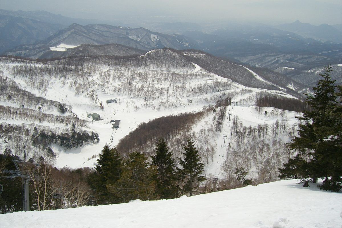 從川場滑雪場上部往下俯瞰：前景是雪坡與針葉樹，下方山谷裡是雪場底部的建物與圓頂小屋，幾條吊椅纜車沿著雪道往上拉，遠方是層層山稜與霧濛濛的平地