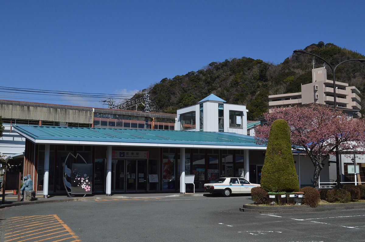 伊豆急行河津駅站舍:藍綠色屋頂,入口上方寫著「河津駅 Kawazu Station」,站前停著一輛計程車,右側一棵開著粉紅花的樹