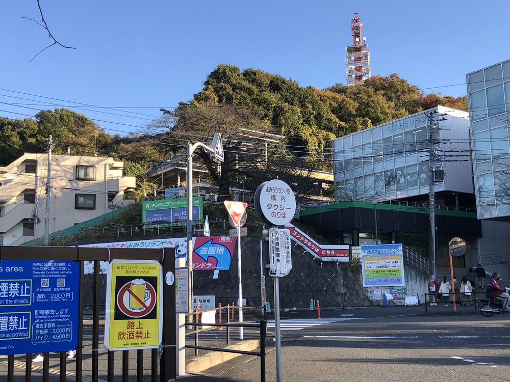 從京王よみうりランド駅前看纜車京王口站:紅色箭頭招牌寫著ゴンドラのりば こちら,後方是玻璃外牆的建築,旁邊有計程車乘車處站牌與纜車票價看板