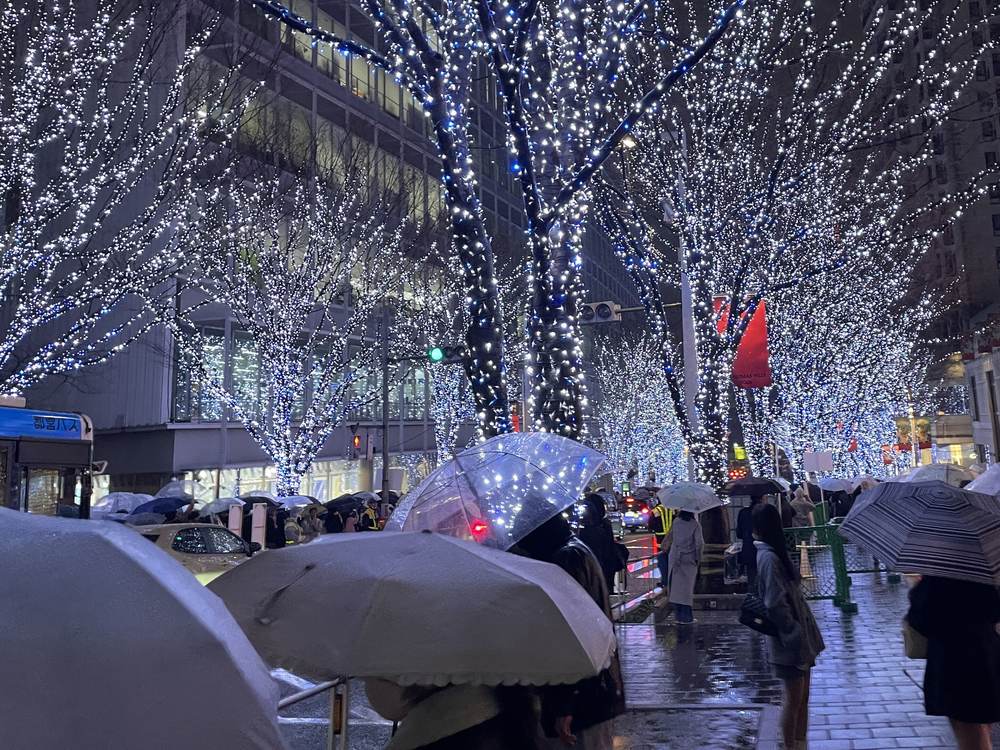 雨夜的六本木けやき坂：行道樹纏滿白色與藍色燈泡，樹下人行道擠滿撐傘的人，左邊停著都營巴士