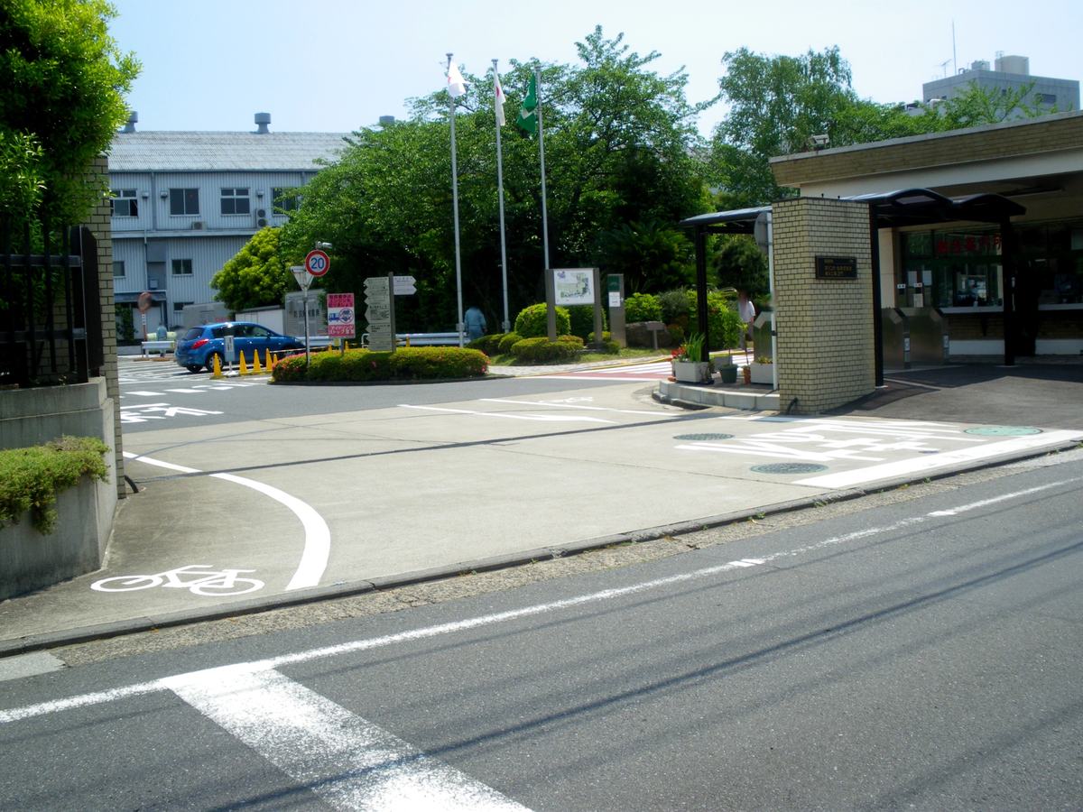麒麟啤酒橫濱工場的車輛出入口:畫面右側是一座淺黃色磚砌的門柱,柱面嵌著一塊深色金屬銘板,上頭印著 KIRIN 標誌與「キリンビール株式會社 橫濱工場 正門」;門柱右邊是一間開著窗的警衛室,玻璃上貼著紅色大字「總合案內所」,窗前排著幾支不鏽鋼的擋車柱與一段黑色雨遮走廊。左側是一條進廠道路,路口立著限速 20 的圓形標誌、幾個指向廠內各設施的箭頭指標牌與三支旗桿,遠處停著一輛藍色小車,路面上畫著白色的通行標線與一個自行車圖示。晴天,樹很綠