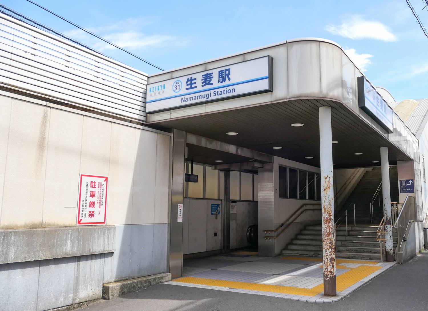 京急線生麥站的東口出入口:一棟灰白色的站體,屋簷下掛著藍白配色的橫式站名燈箱,左端是 KEIKYU 京急電鐵標誌與藍圈裡的車站編號 KK31,中央大字寫著「生麦駅」,下面一行英文 Namamugi Station;出入口是一個沒有閘門的開放式穿堂,裡面看得到一道通往月台的水泥樓梯與扶手、天花板嵌燈,以及地面上的黃色導盲磚,右側牆上另有一塊寫著電梯的指示牌。左邊白色外牆上貼著一張紅框直式告示,大字寫著「駐車厳禁」。晴天,畫面裡沒有人