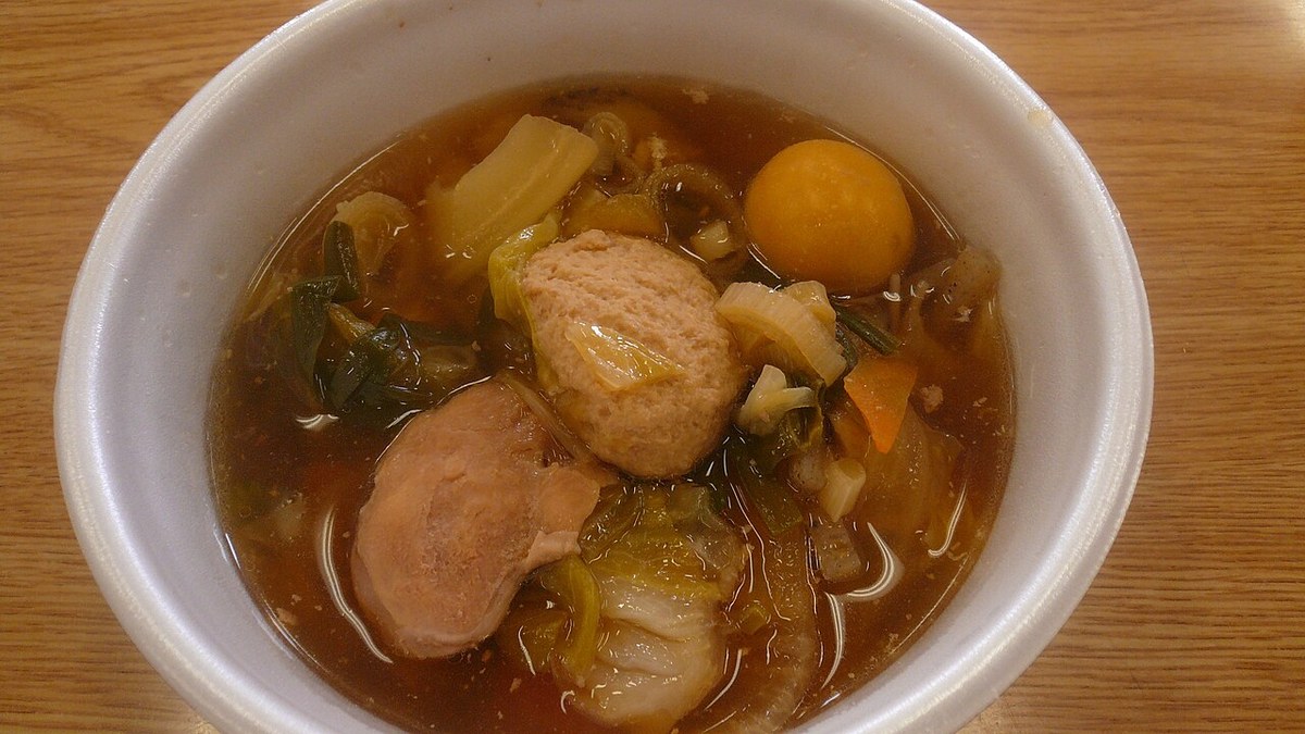 白色碗裡的ちゃんこ鍋特寫：褐色湯裡有一塊雞肉、一顆雞肉丸、白菜、紅蘿蔔、青蔥與一顆橘色圓形食材，碗放在木頭桌面上