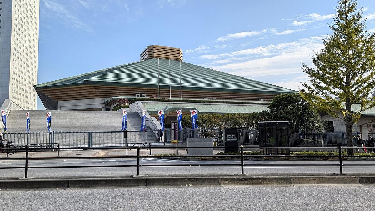 從国技館通り對街拍的両国国技館：綠色四坡屋頂中央有金褐色方形頂塔，白色石牆前立著一排藍色直式旗幟，右側銀杏樹葉已轉黃