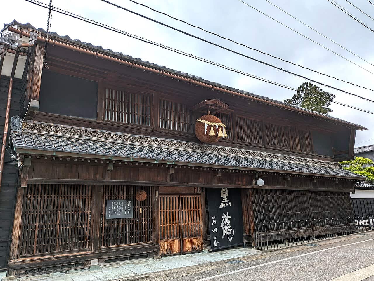 福井縣永平寺町的黑龍酒造店舖,兩層樓深色木造町家,二樓格子窗下的小屋頂中央掛著一顆繫著注連繩的杉玉,一樓入口右側掛著寫有「黒龍 石田屋」的黑色招牌,左側牆上有一塊直書日文的黑色說明板