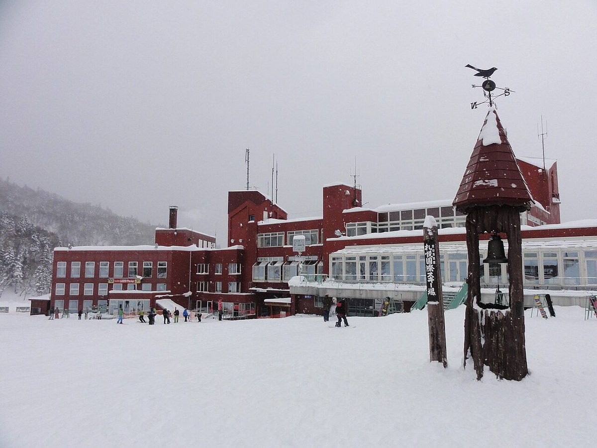 降雪中的札幌国際スキー場 山麓,暗紅色中心建物屋頂積著厚雪,前方緩坡上散布滑雪客,右側木造鐘塔掛著寫有「札幌国際スキー場」的直式看板