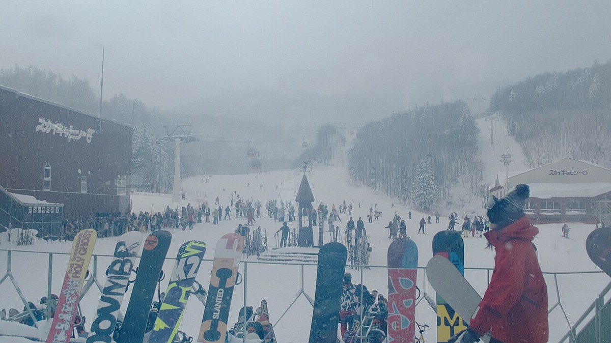 降雪中的札幌国際スキー場 山麓,前景欄杆插滿雪板與雪杖,中景大量滑雪客聚在緩坡上,左右兩棟建物都寫著「スカイキャビン」,廂型纜車沿索道往霧中爬升