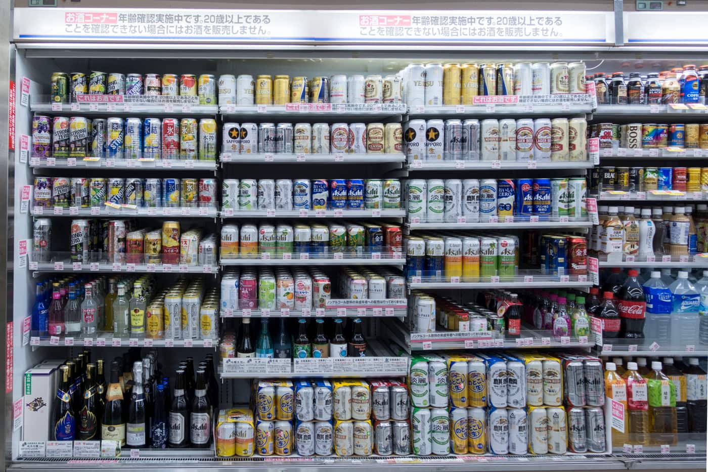日本超商的酒類冷藏櫃全景,上緣紅字橫幅寫著「お酒コーナー」與年齡確認告示,左側整片是罐裝沙瓦,中央與右側是罐裝啤酒、発泡酒與新ジャンル,每一格底下都夾著小張粉紅色價標