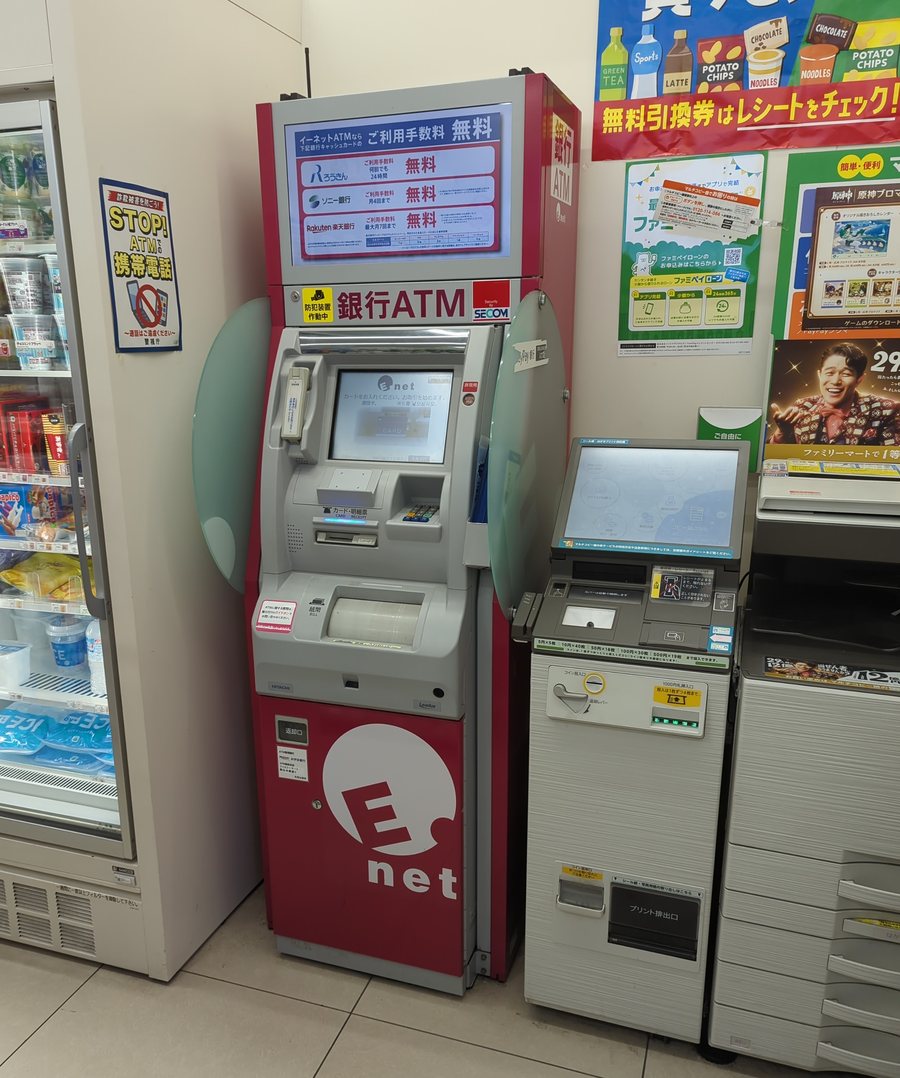 全家超商店內靠牆的紅色 E-net 銀行 ATM，機身印著 E-net 標誌與「返却口」，上方燈箱列著ろうきん、ソニー銀行、楽天銀行三家的手續費免費條件，左邊牆上貼著「STOP! ATM で携帯電話」的防詐海報；ATM 右側並排一台有觸控螢幕、硬幣投入口與「プリント排出口」的多媒體事務機