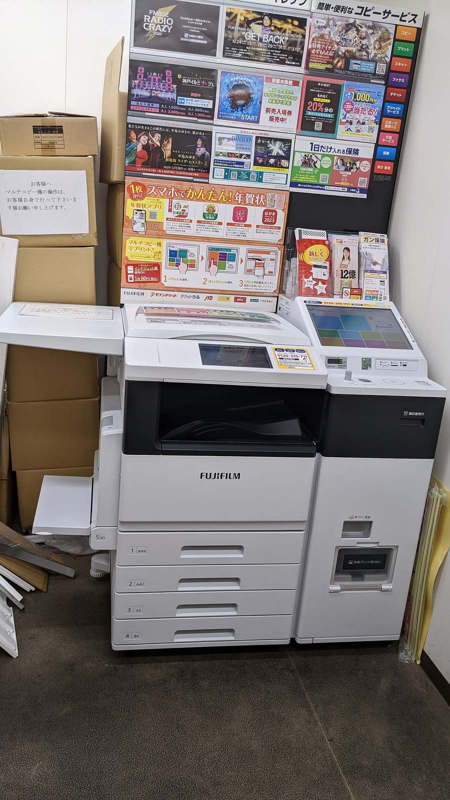 7-ELEVEN 店內角落的白色 FUJIFILM 多媒體事務機，右側連著一台附觸控螢幕與投幣口的付款機；背後牆上是一整面演唱會與展覽的宣傳單，右上角黑底看板直排列著コピー、プリント、スキャン、ファクス、チケット、保険等服務項目；左側疊著紙箱，箱上貼著一張手寫告示