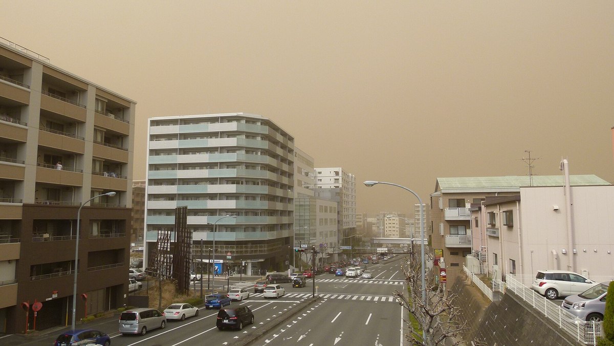 橫濱市區的十字路口與雙向車道，兩側是公寓大樓；整片天空是均勻的土黃褐色，沒有雲的形狀，遠處幾棟大樓的輪廓被霾吃掉、只剩灰白剪影