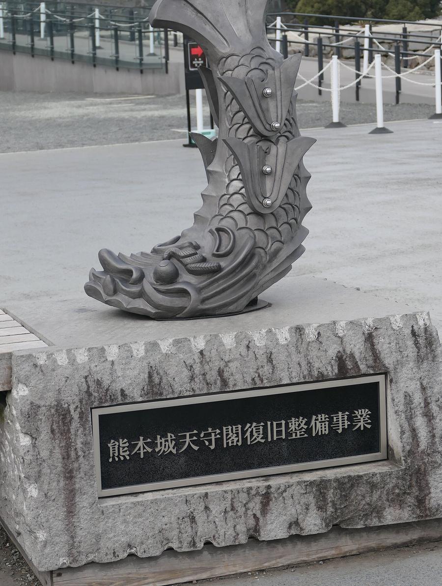 天守閣前廣場上一座實物大的鯱瓦立在石座上,石座正面的黑色銘板刻著「熊本城天守閣復旧整備事業」,背後是特別見学通路的玻璃欄杆