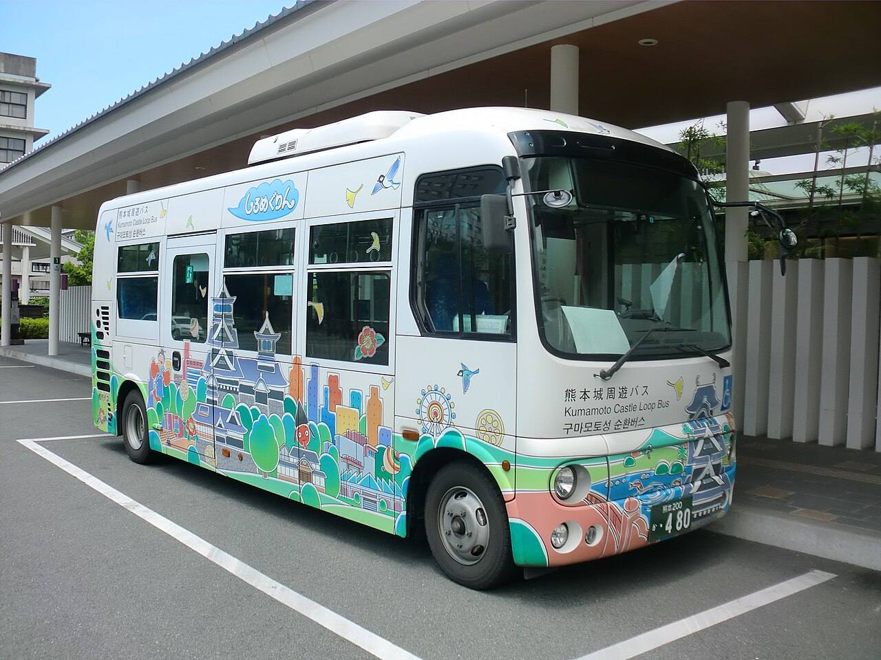 白色小型巴士車身滿是彩色插畫,車頭寫著「熊本城周遊バス/Kumamoto Castle Loop Bus/구마모토성 순환버스」,側面有「しろめぐりん」標誌,停在候車亭旁的停車格
