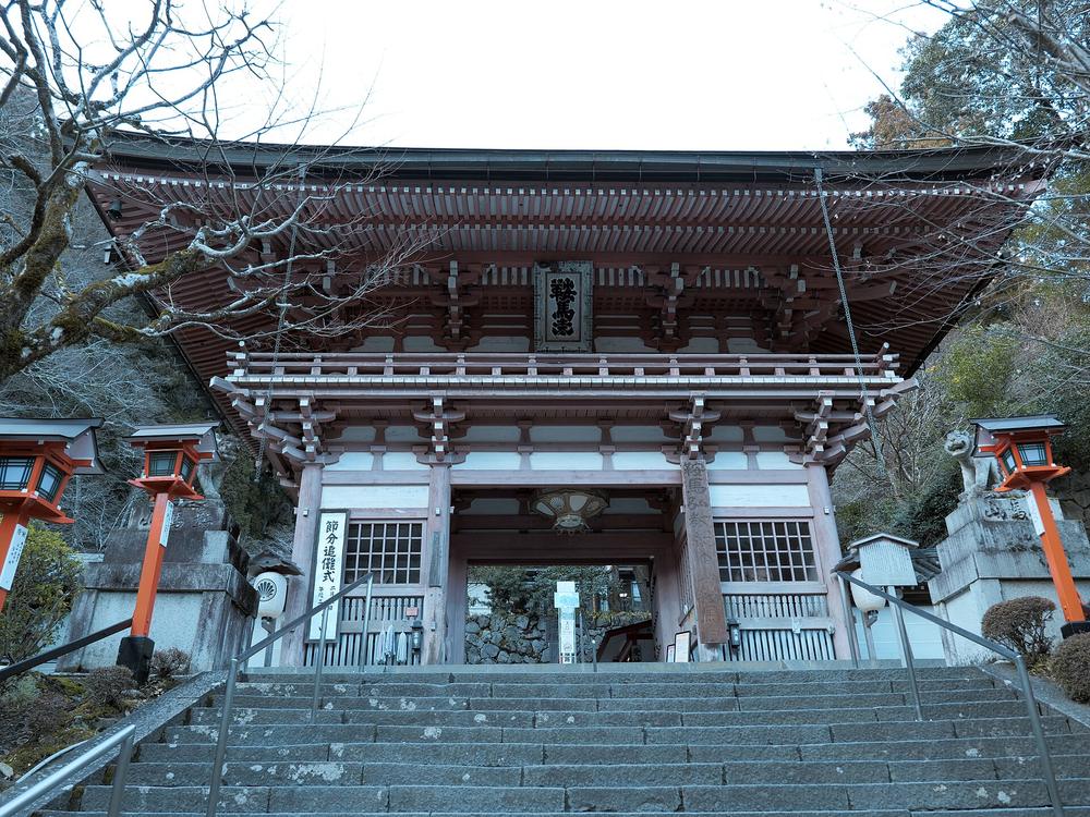 白天從石階下方仰望鞍馬寺仁王門:紅色木造樓門、門上掛「鞍馬山」匾額,門前是一段寬大的石階,兩側有扶手和紅色石燈籠