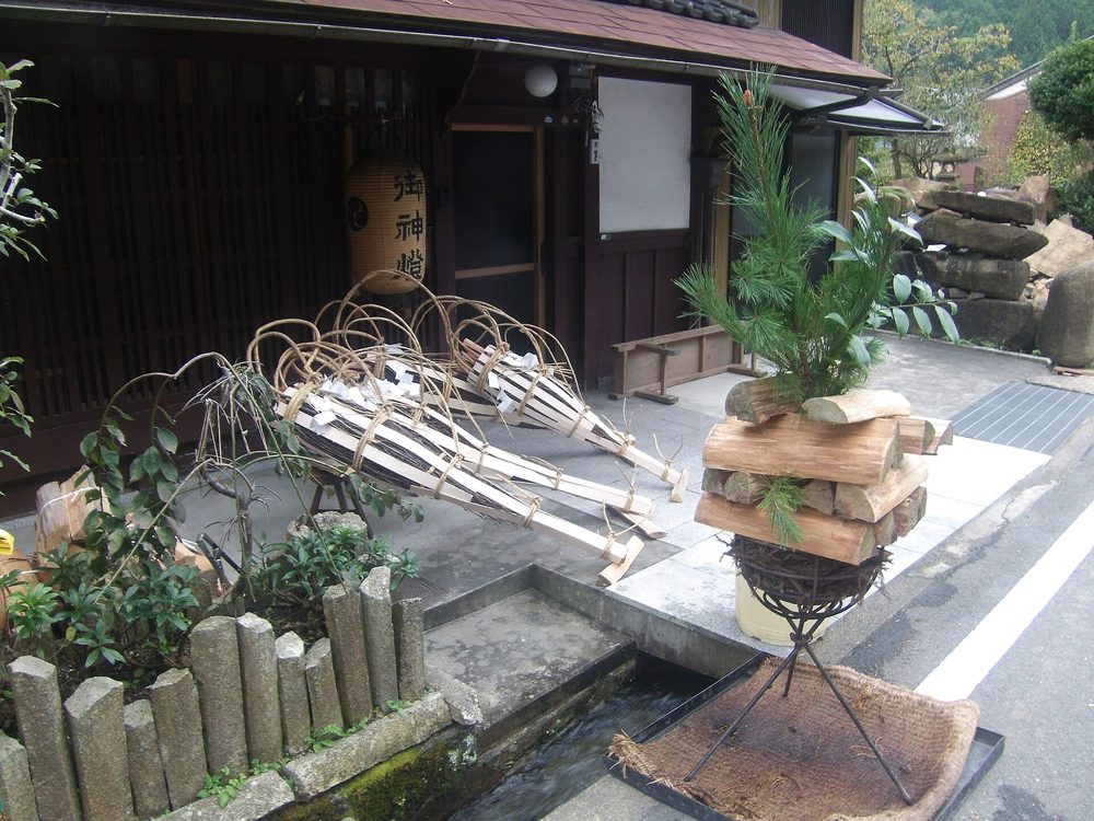 鞍馬一戶民家門前:鐵架上堆好一疊劈開的木柴、頂上插著松枝,左邊地上擺著三支綁繩圈的松明,屋前有小水溝,門邊掛「御神燈」燈籠