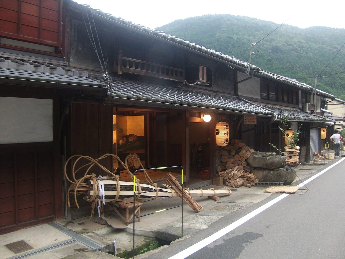 火祭當天傍晚前的鞍馬街道:左邊一排木造老屋,門口擺著綁好繩圈的大松明和堆起的木柴,屋簷下掛「御神燈」燈籠,右邊是空著的柏油車道,背後是綠色山坡
