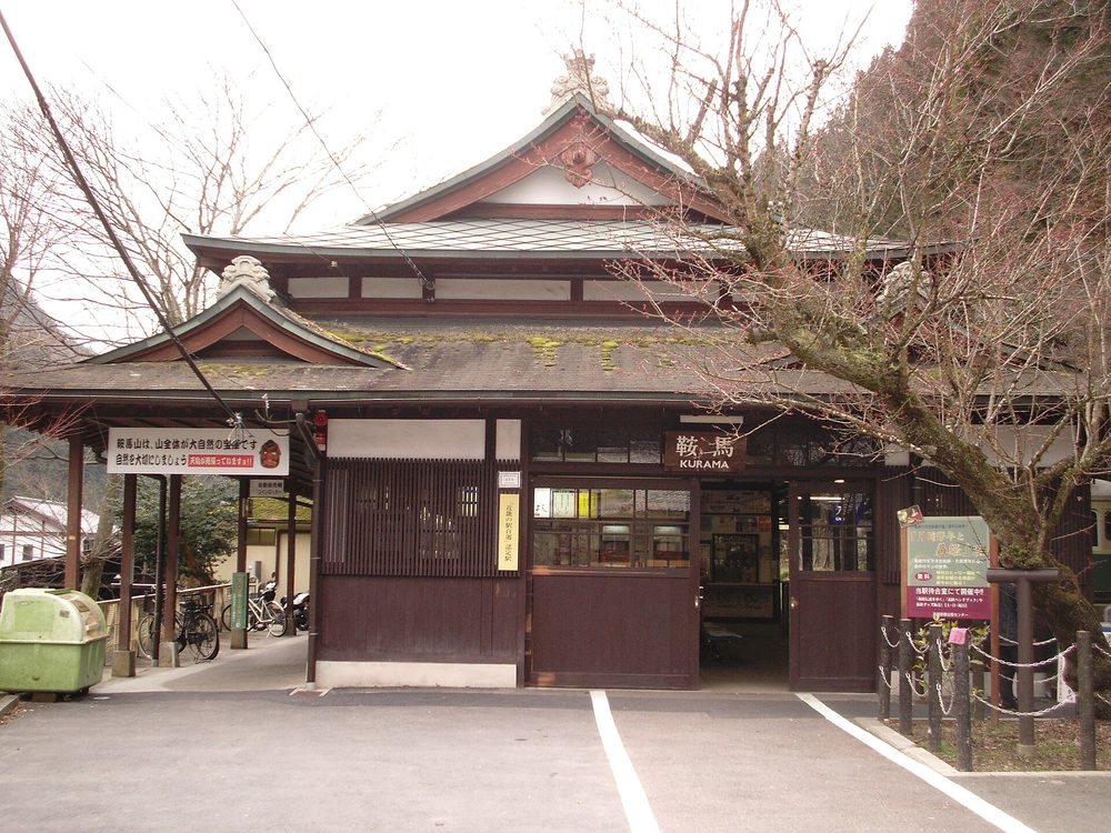 叡山電車鞍馬站的站房正面:單層木造建築、瓦頂上有山形破風,入口上方掛「鞍馬 KURAMA」木牌,站前是畫白線的空地,右邊一棵枝葉光禿的樹
