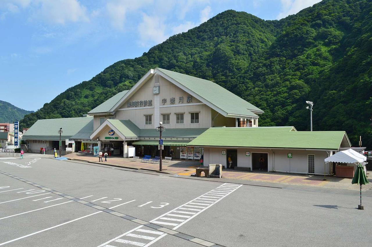 黑部峽谷鐵道宇奈月站站舍,淺綠色屋頂的山形建築,山牆上有時鐘與站名,前方是空曠的大型停車場