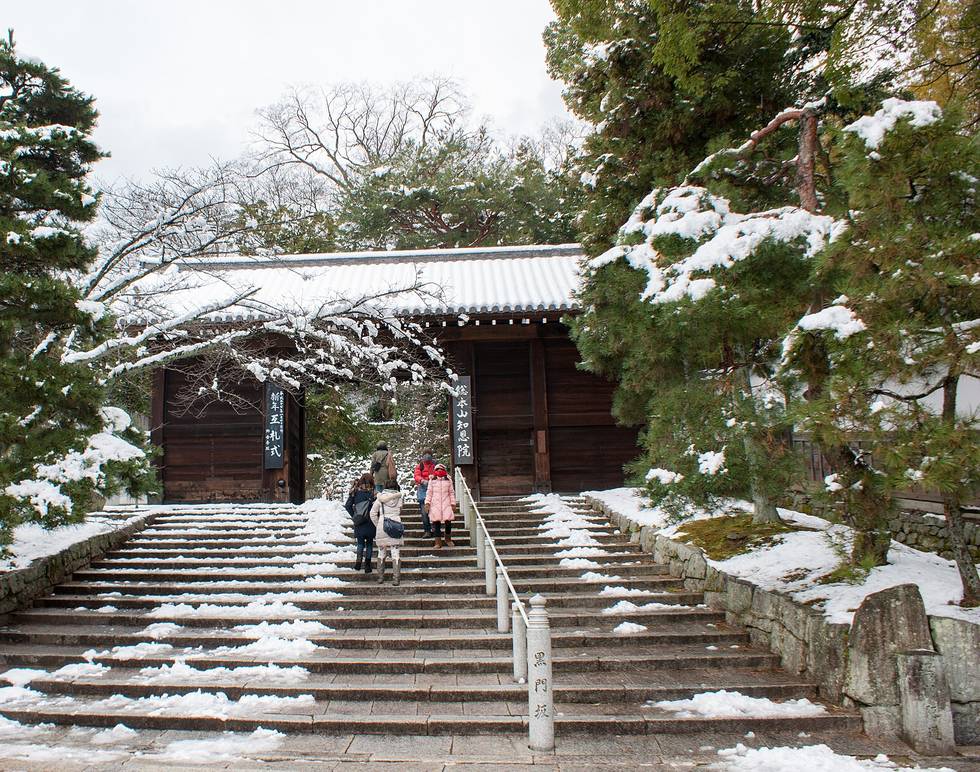 知恩院黑門前的石階：雪已經融掉一半，只在每一階的邊緣與兩側石壁上留下一條條白色殘雪，石面是濕的、露出深色，門頂與一旁的松樹還積著雪，幾個背對鏡頭的遊客正往上走