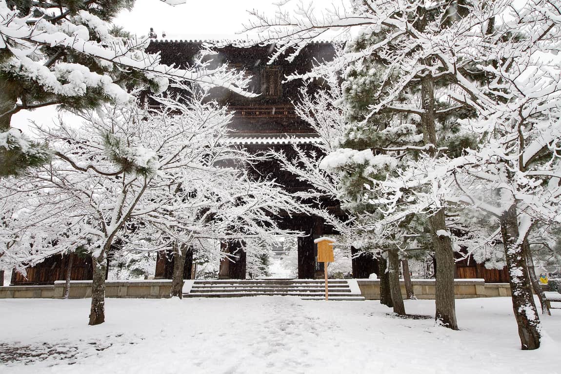 大雪中的兩層寺院山門,屋頂與周圍的松樹、櫻樹枝條全積著雪,門前石階與整片地面覆雪,只有一行腳印,門旁立著一塊木製告示牌,畫面裡沒有半個人