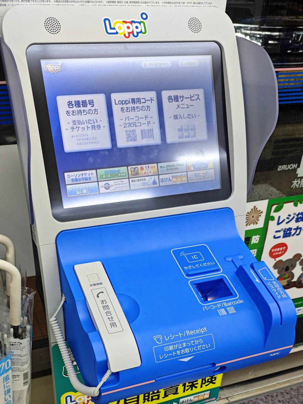 LAWSON 店內的白藍色 Loppi 機台正面：上方是觸控螢幕，首頁橫排三個按鈕，左起「各種番号をお持ちの方」「Loppi 専用コードをお持ちの方」「各種サービスメニュー」；下方藍色斜面上有 IC 感應區、條碼掃描窗、左側一具捲線話筒標著「お問合せ用」，最前緣印著「レシート／Receipt 印刷が止まってからレシートをお取りください」