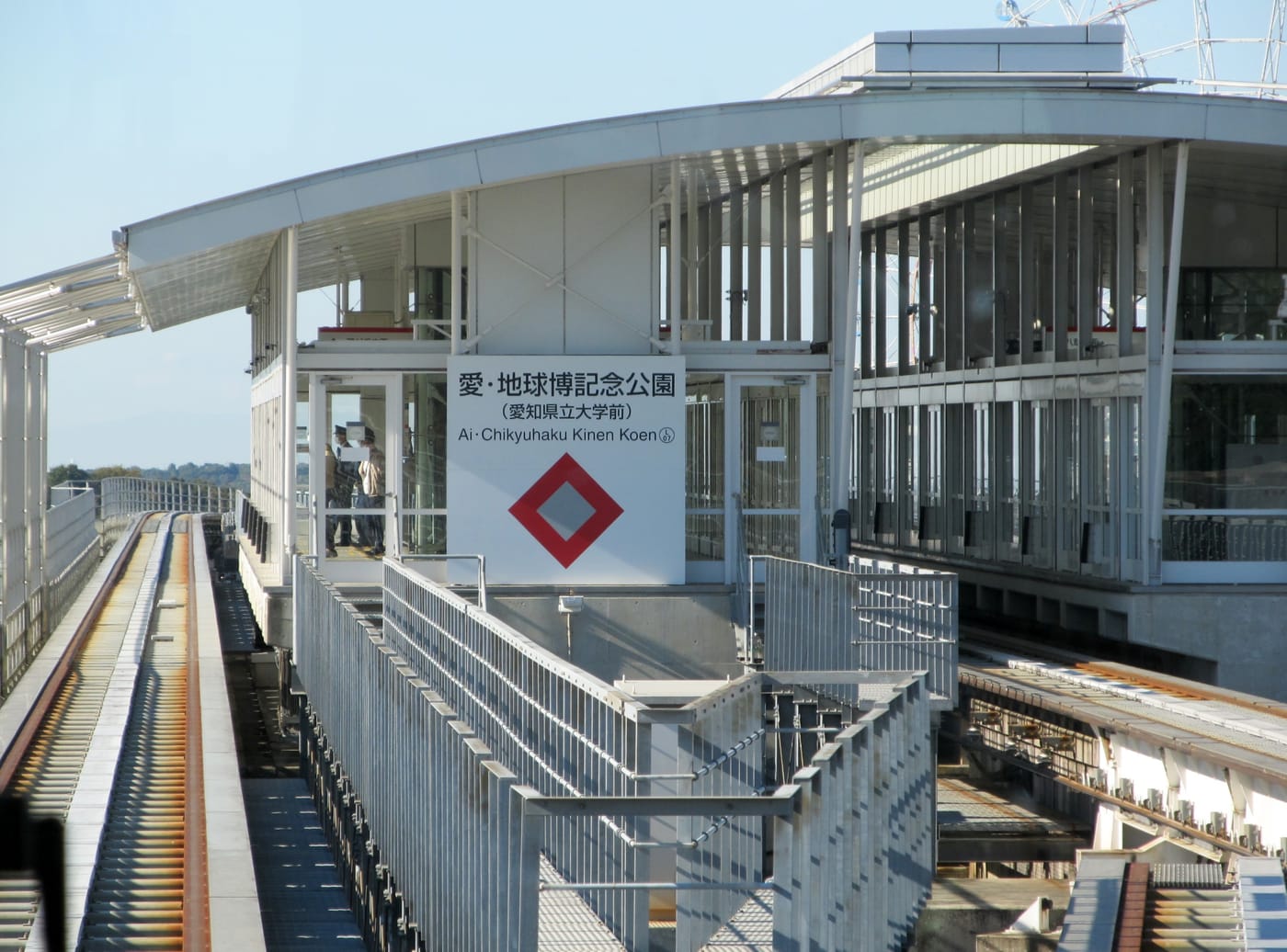 Linimo 磁浮愛・地球博記念公園駅的高架月台，白色站名牌寫著「愛・地球博記念公園（愛知県立大学前）Ai･Chikyuhaku Kinen Koen」與一個紅色菱形標誌，左側是空無列車的高架軌道