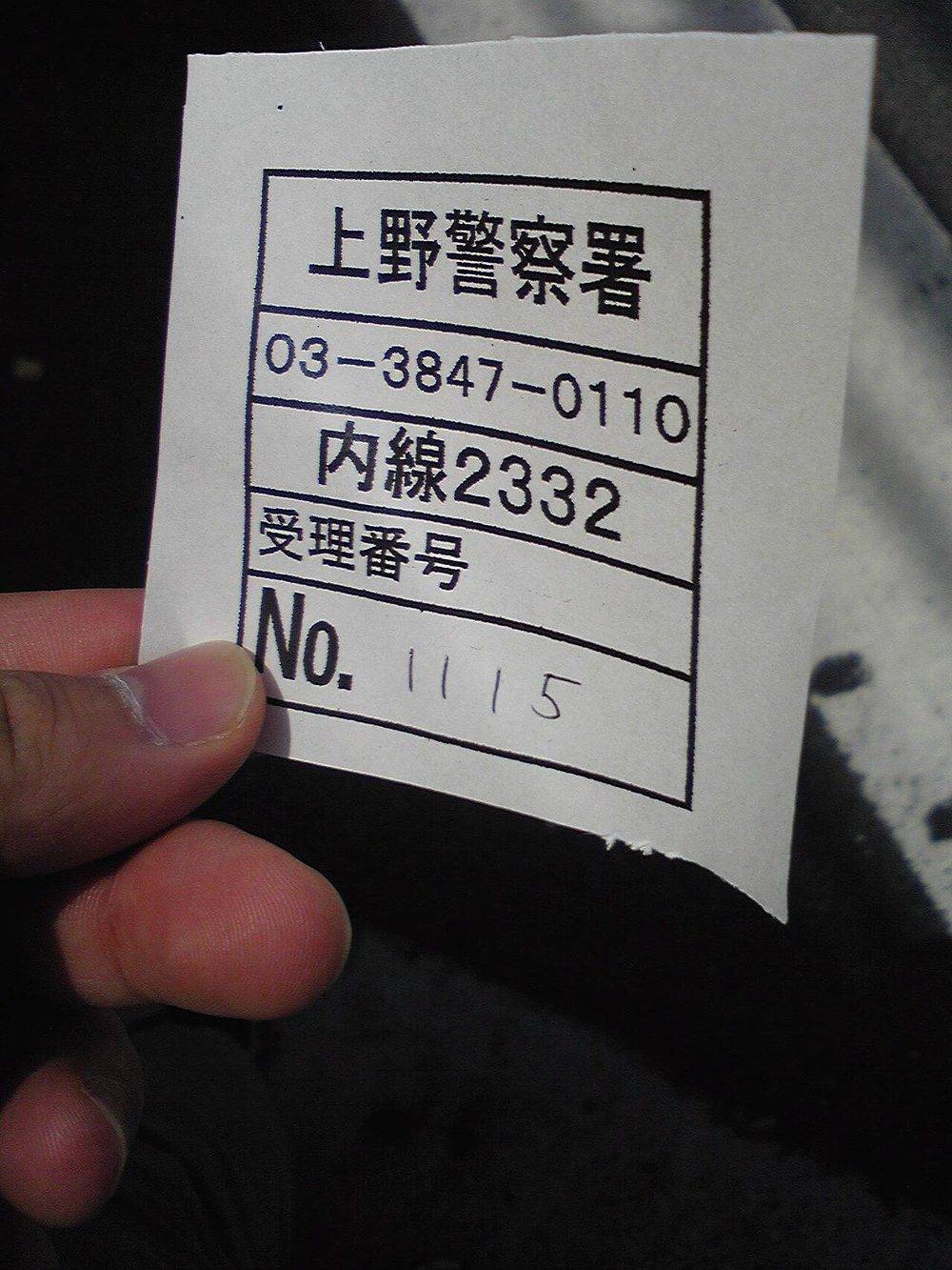 一隻手捏著一張撕下來的白色小紙片,上面印著方框表格,由上而下依序是粗體的「上野警察署」、電話「03-3847-0110」、「内線2332」、「受理番号」,最後一格印著 No. 並用原子筆手寫著 1115