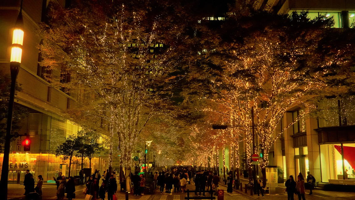 丸之內仲通夜景，兩排行道樹纏滿暖金色 LED 燈串形成光隧道，人行道上是穿大衣的行人與兩側辦公大樓
