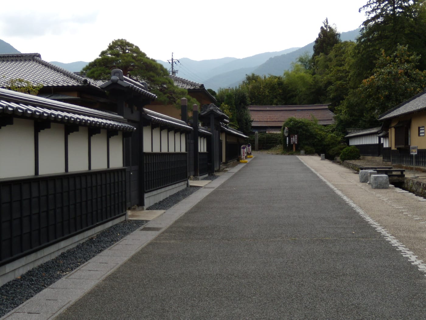 松代町的舊武家屋敷街道:左側一整排白牆配黑色木格的長屋牆沿路延伸,中央是寬闊的鋪裝路面,右側有石凳與水渠,正前方盡頭是一棟深褐色斜屋頂的門樓,再往後是層層疊起的山稜