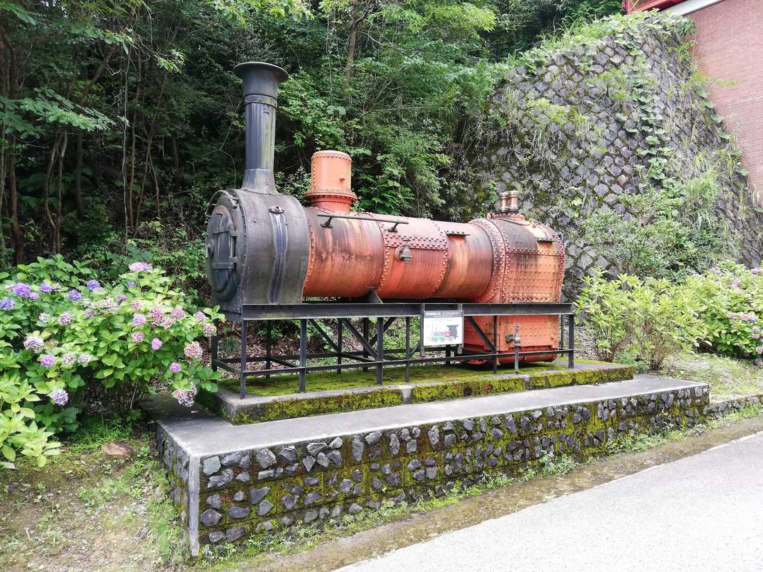 戶外鐵架上橫放著一具鏽紅色的舊式蒸汽機關車鍋爐，前端接黑色煙室與煙囪，鐵架前掛著一塊解說牌，兩側開滿繡球花