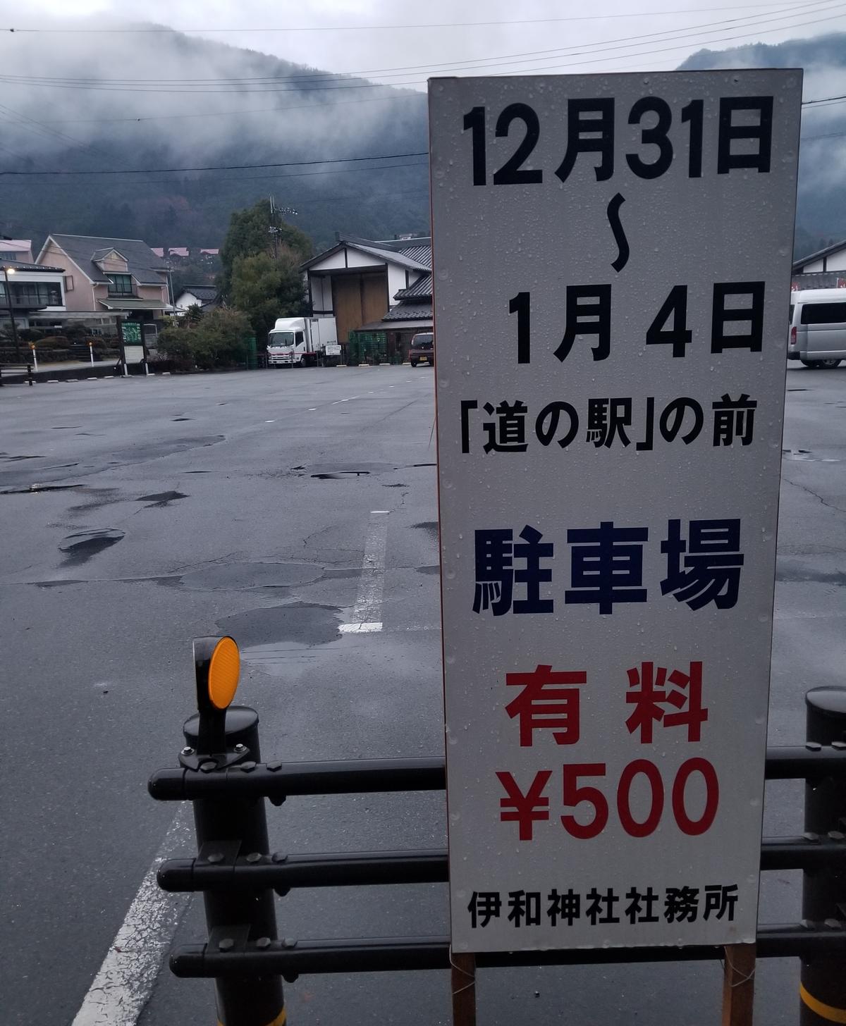 立在道の駅停車場邊的白色手寫立牌,逐字寫著「12月31日〜1月4日 『道の駅』の前 駐車場 有料 ¥500 伊和神社社務所」,後方是雨後空蕩的柏油停車場、護欄與雲霧繚繞的山。