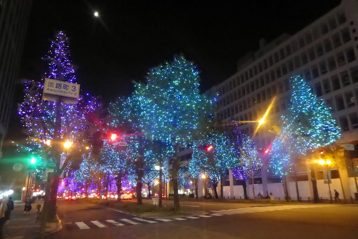 御堂筋淡路町三丁目路口的夜景，路邊一排行道樹整棵纏滿藍色和紫色燈串，左邊掛著「淡路町 3」路牌