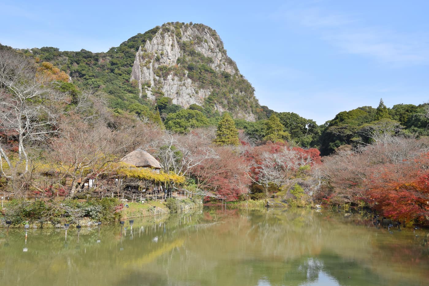 御船山樂園紅葉季白天景致,陡峭岩壁、鏡池倒影與湖畔茅草涼亭旁的紅葉,佐賀縣武雄市