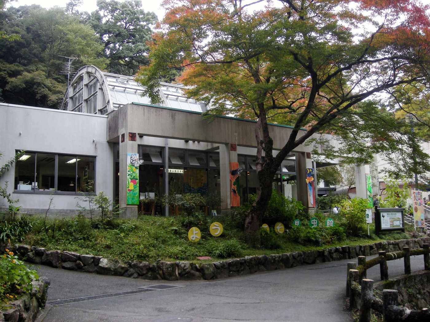 箕面公園昆蟲館外觀,混凝土館舍後方是弧形玻璃放蝶溫室,門前草坡排著平假名字牌