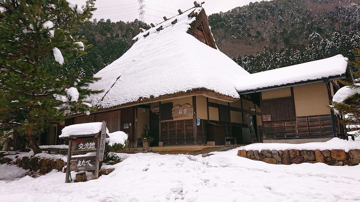 美山かやぶきの里的茅草屋：陡斜的茅草屋頂戴著一頂厚雪帽，門口掛著寫有「彩花」的木牌，庭前立著「交流館　またべ」的木製指示牌，地面的積雪上有腳印
