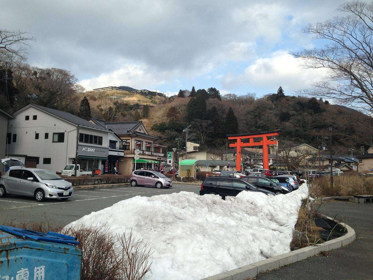 冬季的元箱根路旁,一座朱紅色鳥居立在道路另一側的樹林邊,前方是柏油停車場,車格裡停滿轎車,畫面中央堆著一座被鏟到一起的殘雪堆,左側是兩層樓的商店與蕎麥屋,門口掛著綠色的「P」停車場指示牌,背景是落葉後的褐色山坡與陰天