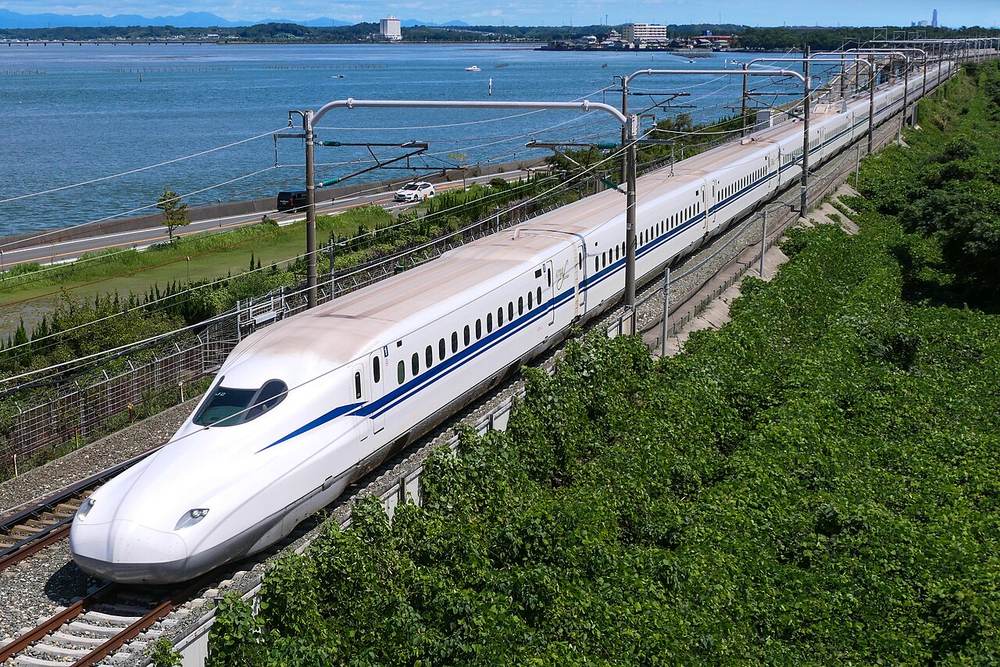 從高處俯瞰的東海道新幹線 N700S，白色車身配一道藍線，沿著水岸邊的路堤畫出弧線駛來，車身往右上角一路延伸到看不見盡頭；對岸有一條公路與停著的兩台車，前景是整片綠色植被與架線柱