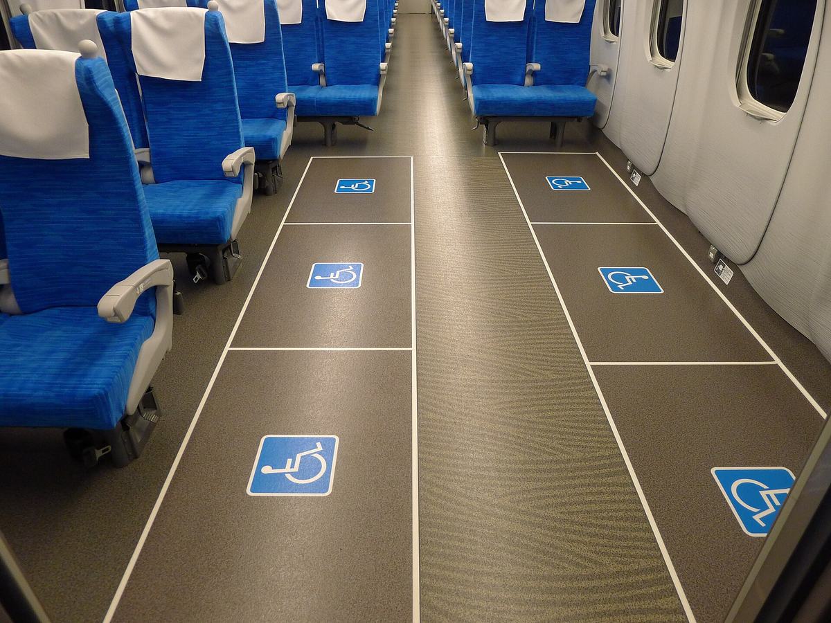 東海道新幹線 N700S 改裝後的車廂內,走道兩側各有三格白線框出的空地板,每格印一個藍底白色輪椅標誌,共六格;靠窗那一列的車壁底部有固定輪椅用的金屬扣件與小型說明牌,另一列旁邊是整排藍色座椅