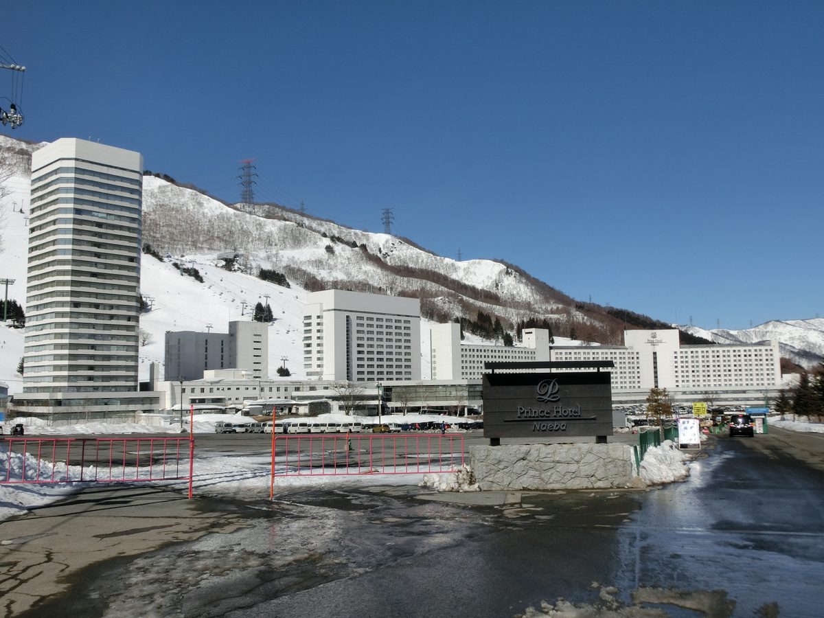 苗場王子飯店入口一帶：前景是融雪潮濕的停車場和粉紅色柵欄，右邊立著深色的「Prince Hotel Naeba」招牌，後方是一棟高塔樓和一整排相連的白色飯店大樓，再後面是有吊椅纜車和電塔的雪山