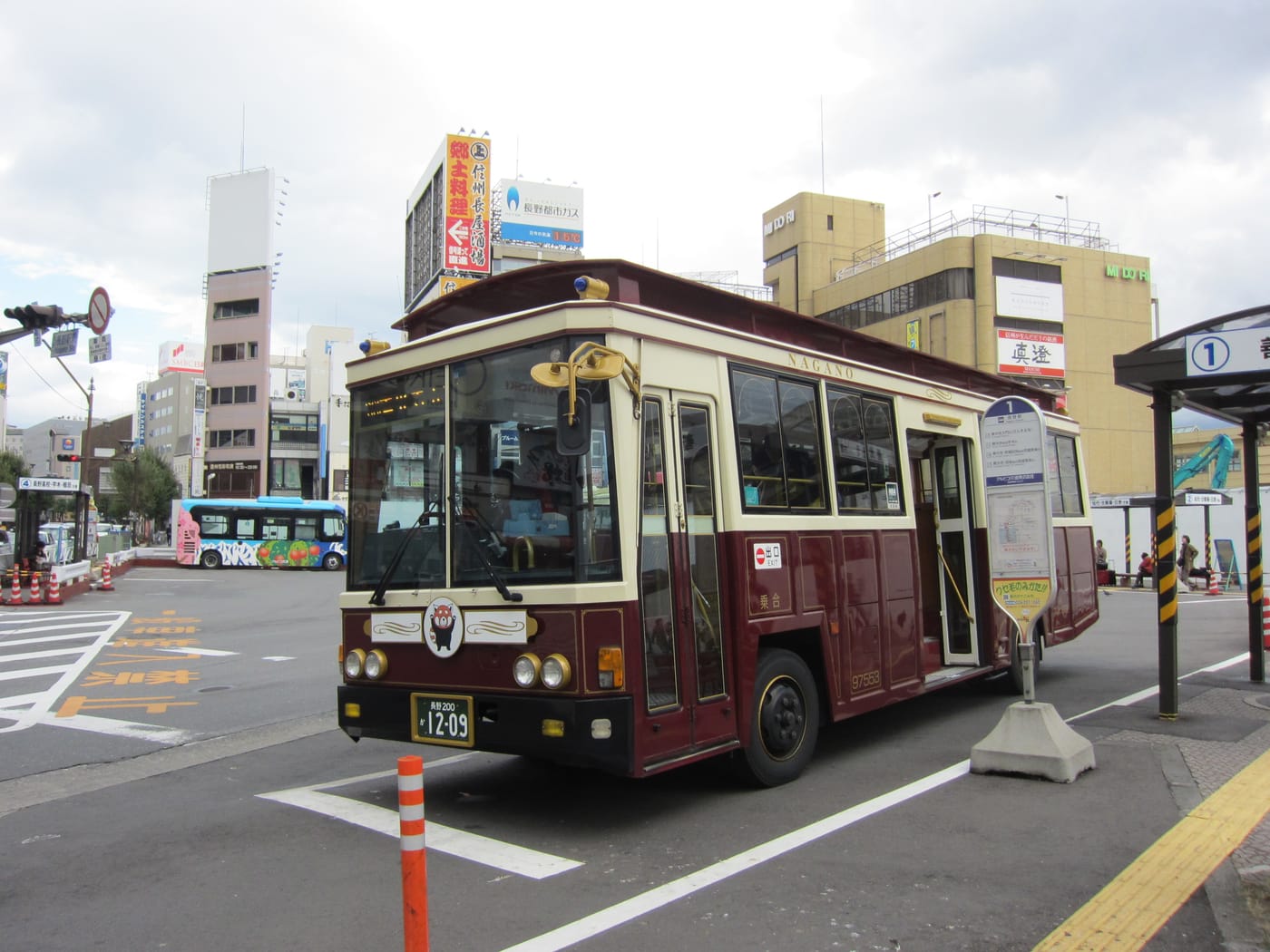 一輛深酒紅配米白車身的復古造型小巴停在長野駅前的巴士乘車處,車頭上方寫著 NAGANO、正面掛著一個白底的圓形吉祥物標誌,右側立著寫有大大「1」字的圓形站牌,站牌上貼著路線時刻表