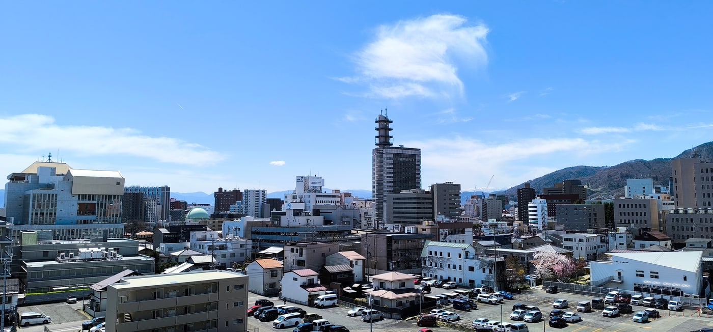 從高處俯瞰長野市中心的四月街景:低矮的住宅與辦公樓一路鋪到山腳,正中央一棟深色高樓頂著白色鐵塔,右側是低緩的山丘,右下角一株櫻花正開在停車場旁邊