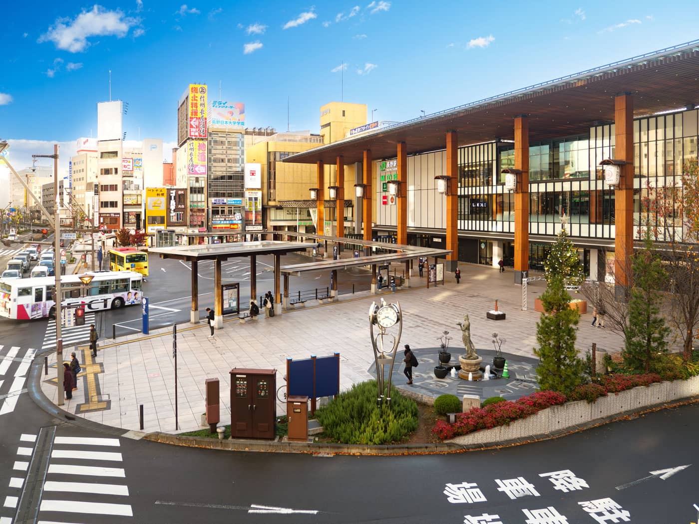 長野駅善光寺口的站前廣場:右側是玻璃帷幕加一排粗大木柱與吊燈的車站正面,中央是有頂棚的巴士乘車島與圓環車道,左側是掛滿直式招牌的市街大樓,廣場中間有一座塔式時鐘與一尊銅像