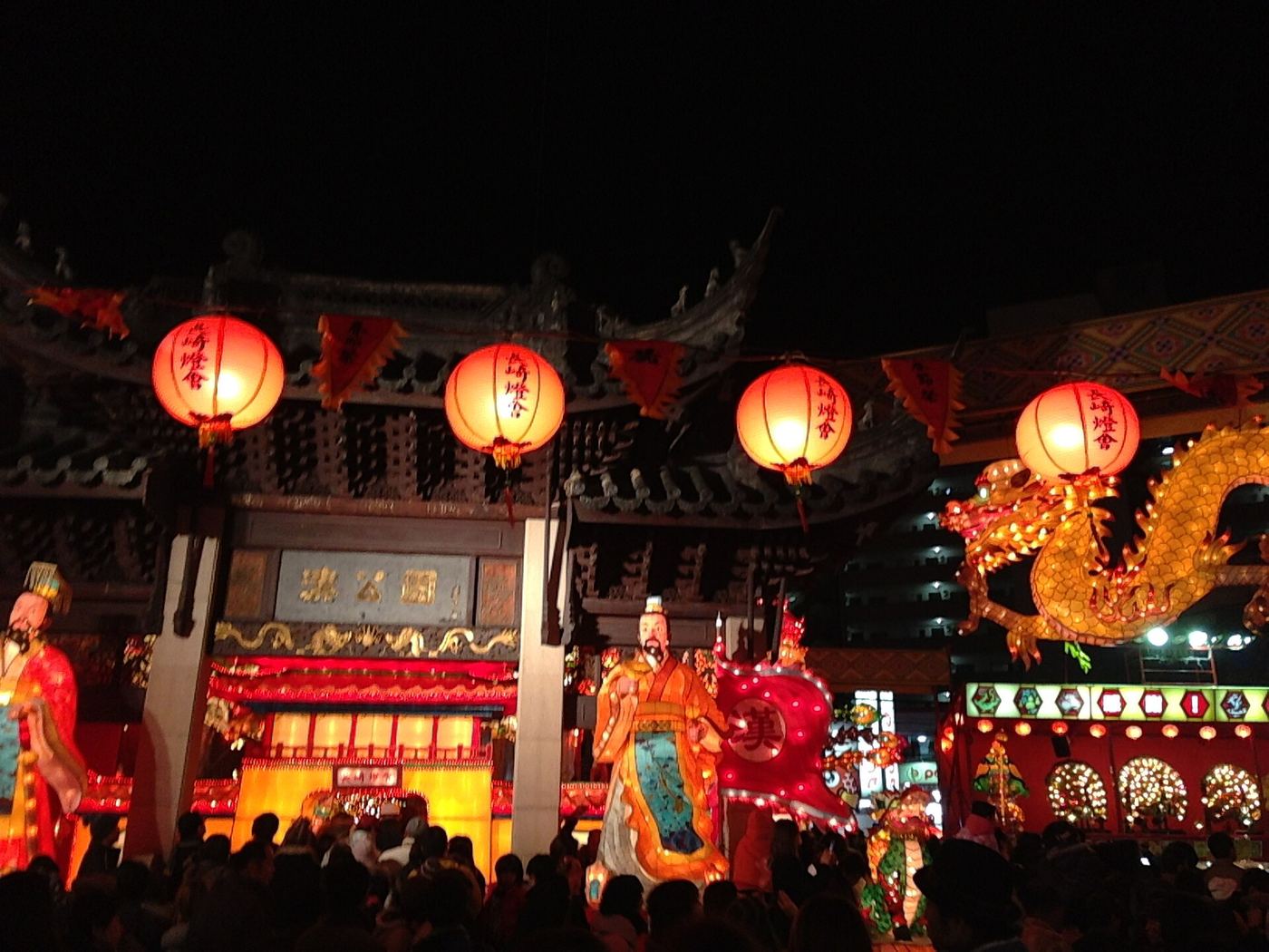 長崎燈會湊公園會場夜景：中式牌樓上掛著四盞寫有「長崎燈會」的大紅燈籠，牌樓匾額刻著「湊公園」三字，右側是一條金色巨龍燈籠，中央立著一尊手持「漢」字旗的古裝人物造型燈籠，前方擠滿人潮