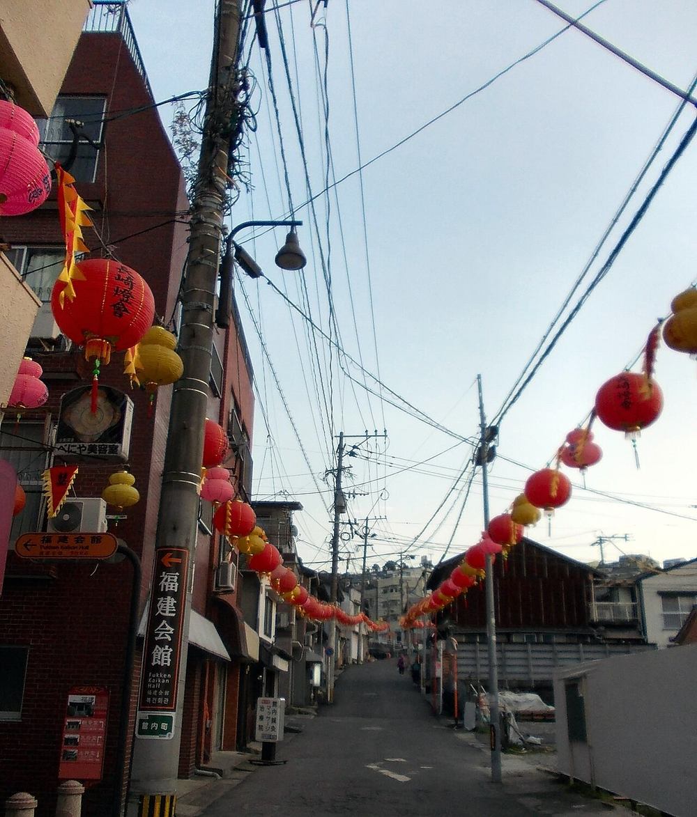 唐人屋敷一帶的窄坡道：兩側是紅磚公寓與老舊木造民宅，電線交錯的天空下拉著兩串紅、粉、黃三色紙燈籠與紅黃三角旗，左側立著寫有「福建会館」的綠色路標，坡道盡頭只有一名行人