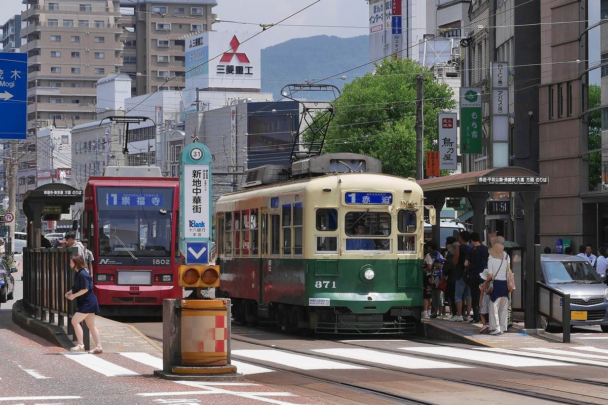 長崎路面電車「新地中華街」停留場：一輛綠色車身、米色車頂的舊型電車停在站台旁，正面方向幕寫著「1 赤迫」，左邊是一輛紅色公車，站牌柱上標著編號 31 與「新地中華街」，月台上有旅客等車