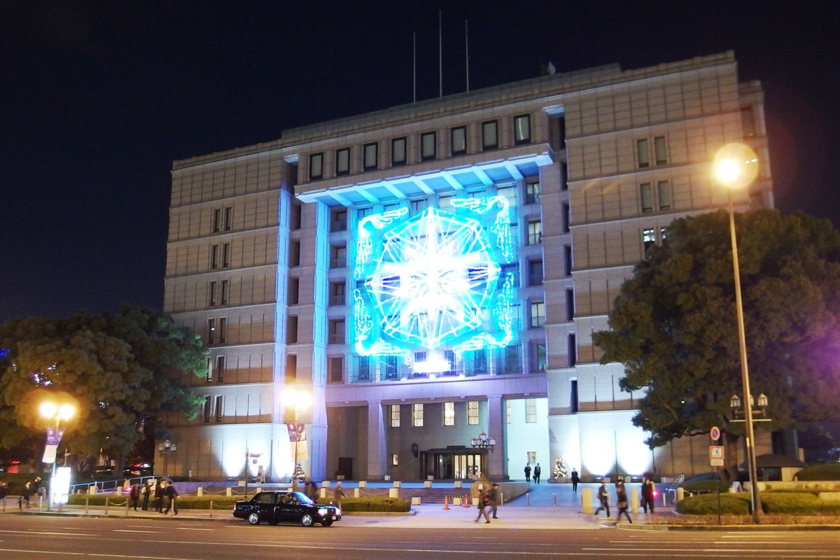 大阪市役所正面夜景，建物立面掛上大型光雕圖騰、樓身被藍白燈打亮，前方是御堂筋車道