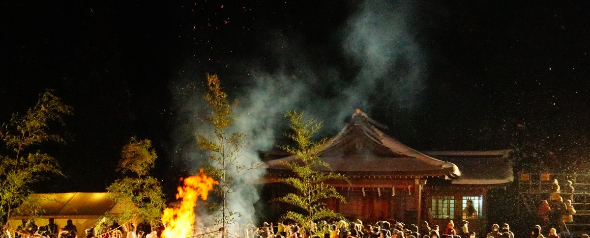 夜間的真山神社境內,一堆柴火在露天燒著,橘紅色的火舌約有一人高,白色的火星從火焰頂端往上飄散;火堆右後方是木造的神社社殿,屋簷下亮著燈,一縷淡青色的煙從火堆斜斜升上夜空,畫面裡佈滿正在下的雪花,畫面最下緣是一整排戴著毛帽與連帽外套、背對鏡頭的觀眾頭頂