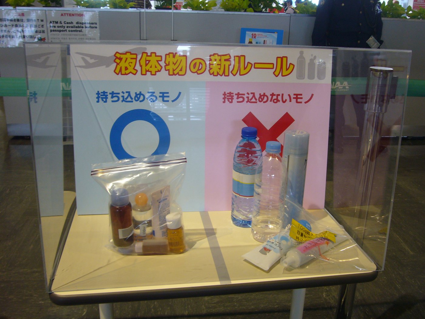 成田機場的壓克力展示箱,背板左半藍底寫「持ち込めるモノ」配一個大大的藍色圈,右半粉紅底寫「持ち込めないモノ」配紅色叉;左側桌面擺著一個裝了數個小瓶的透明夾鏈袋,右側擺著一瓶礦泉水、一支透明塑膠瓶、一罐細長噴霧罐,前方兩條牙膏上貼著黃色標籤