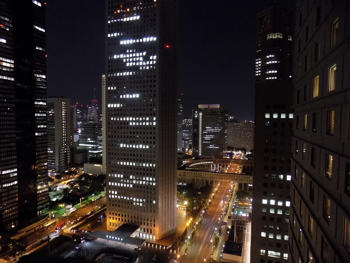 從高樓俯瞰西新宿夜景,中央一棟白色摩天樓亮著格狀窗光,左右兩側是深色高層建築,中間馬路串著車燈與路燈,並可看到一段弧形人行陸橋